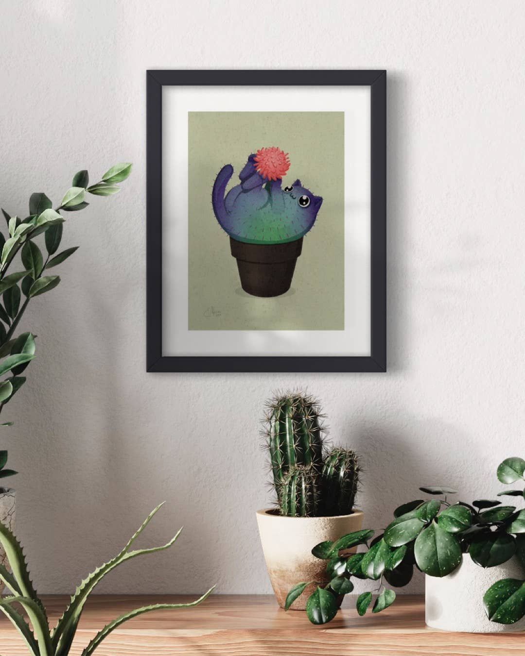 The Coral Prickly Purr Catcus Art Print (7 x 5) by Elissa Marie Creative, featuring a smiling kitty cactus with a pink flower, hangs on a white wall above two potted plants: a real cactus and a leafy plant.