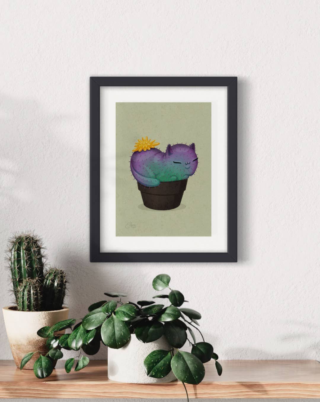 The Yellow Prickly Purr Catcus 7x5 art print by Elissa Marie Creative decorates a white wall above a wooden surface with two potted plants, including a cactus and a leafy plant.