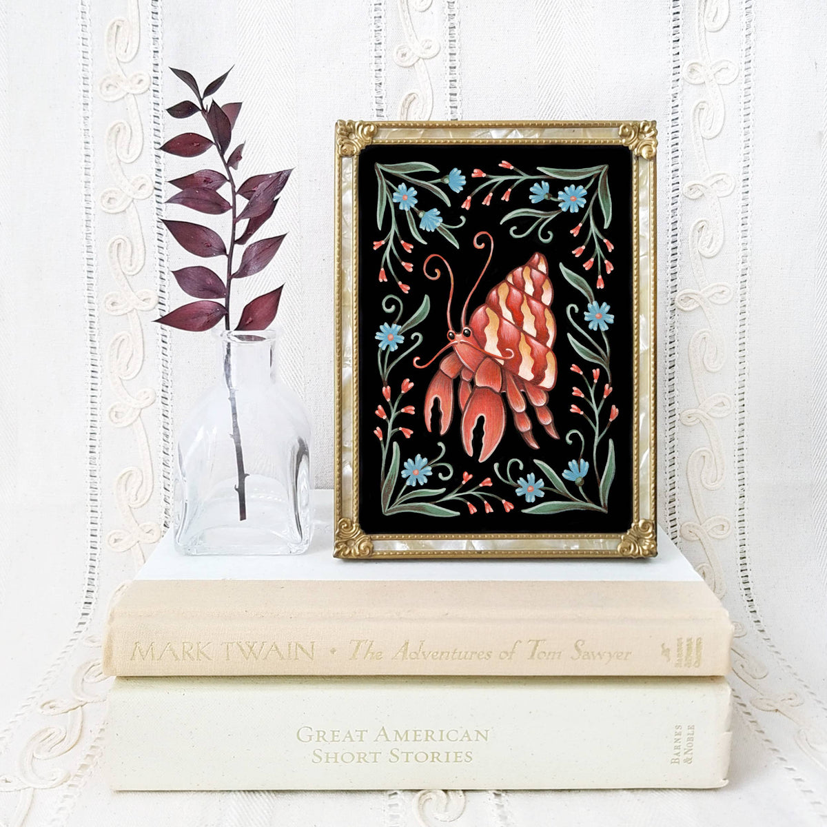 A framed Faina Lorah *Retiring* Crab Folk Art Print in various sizes sits on a stack of Mark Twain books, next to a glass vase filled with dark burgundy leaves.
