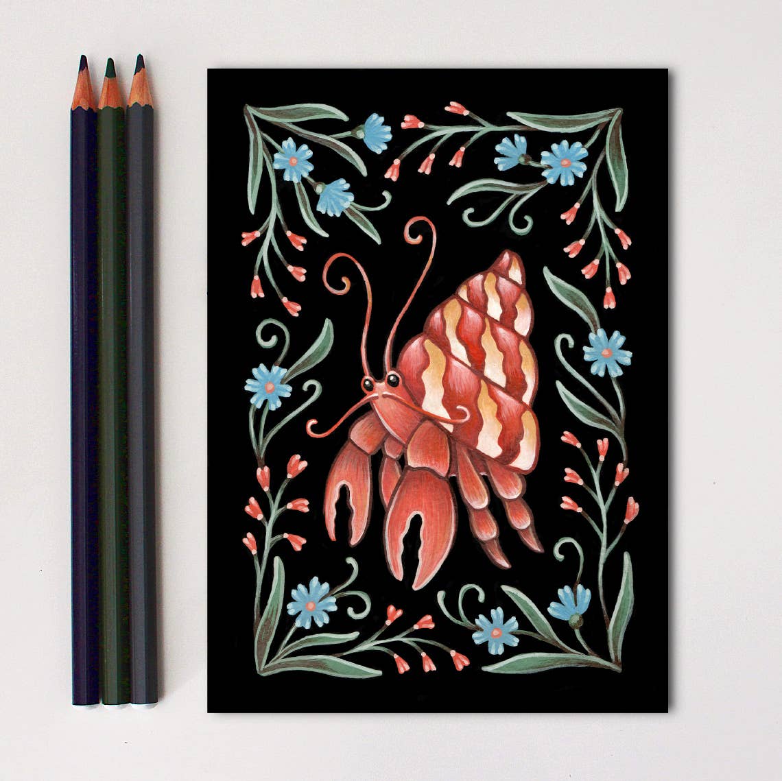 The *Retiring* Crab Folk Art Print by Faina Lorah features a hermit crab with a spiral shell amid blue flowers and green leaves on black. Blue, green, and brown pencils sit to the left. Inspired by the giclée method; available in multiple sizes.