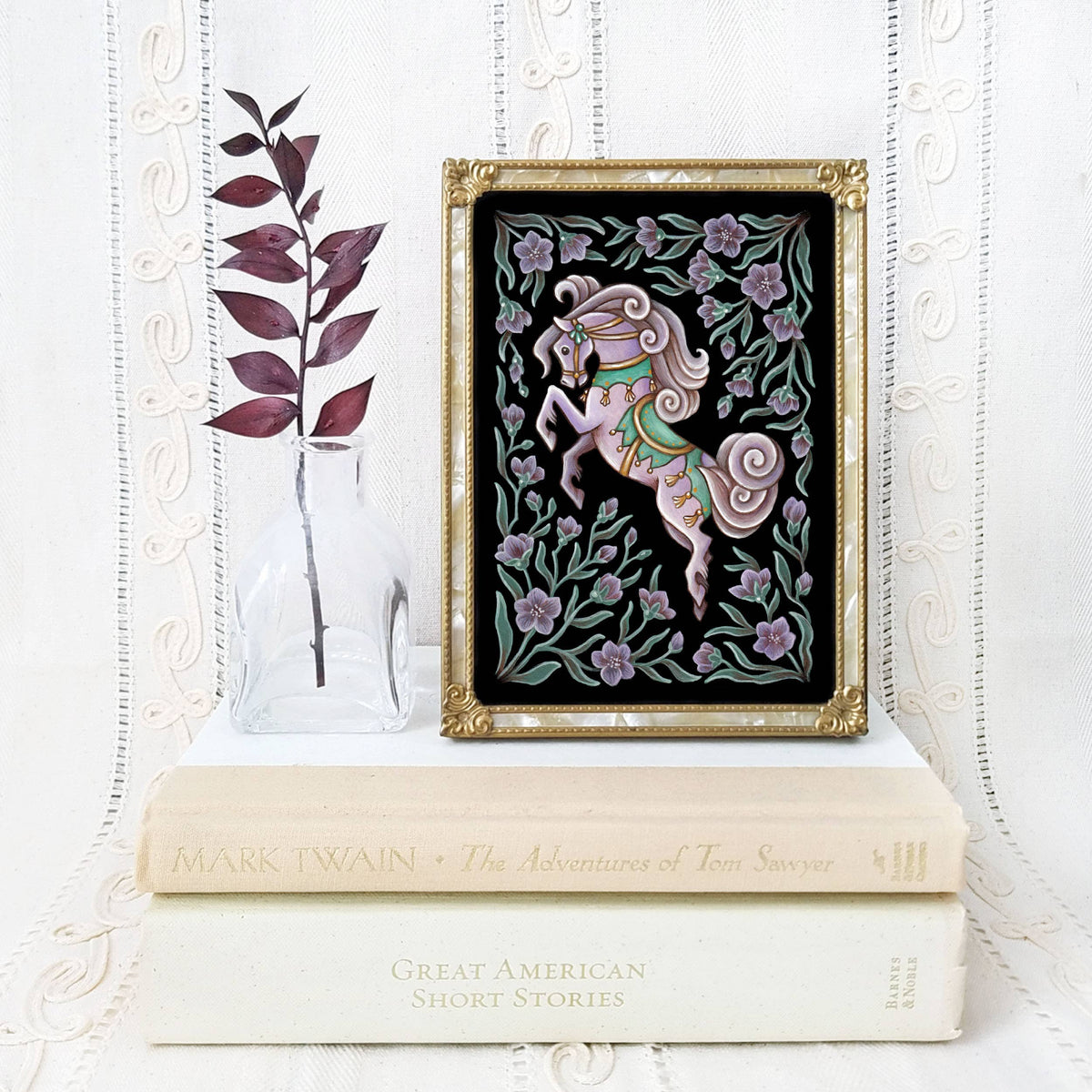 The *Retiring* Horse Folk Art Print by Faina Lorah, featuring a pink carousel horse with flowers, is displayed above classic books alongside a glass vase and dried branch, set against a white lace background.