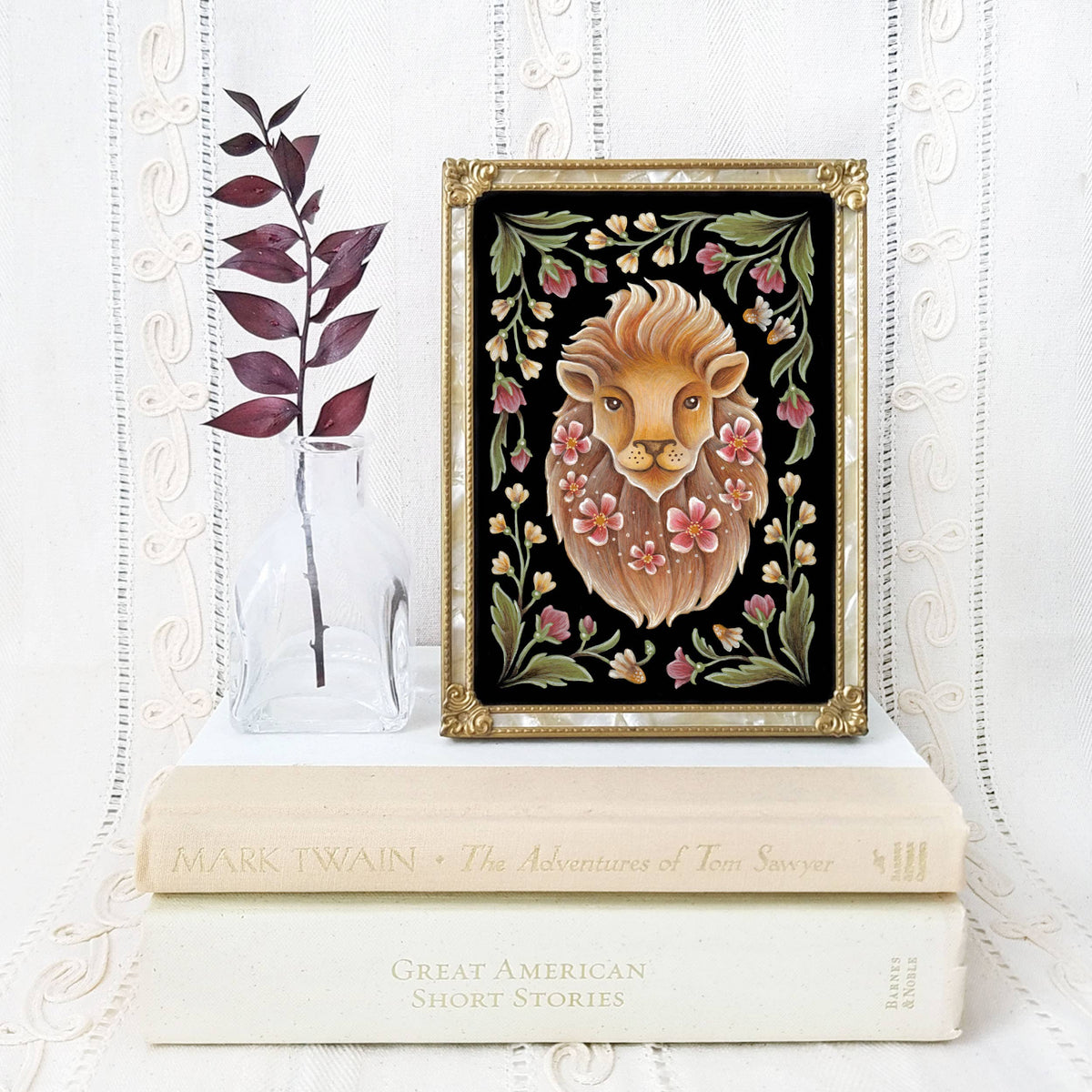 The *Retiring* Lion Folk Art Print by Faina Lorah, available in multiple sizes, features a watercolor lion among pink flowers and green leaves. Shown framed atop books beside a clear vase with a dried branch against a light textured background.