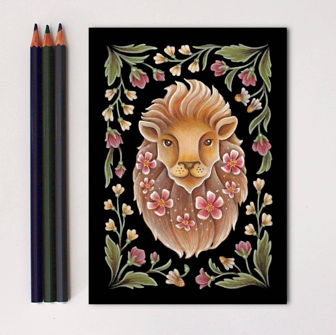 The *Retiring* Lion Folk Art Print by Faina Lorah features a lion’s face with a flowing mane, pink flowers, and green leaves on black. Three colored pencils sit to the left of this vibrant artwork, available in multiple sizes.