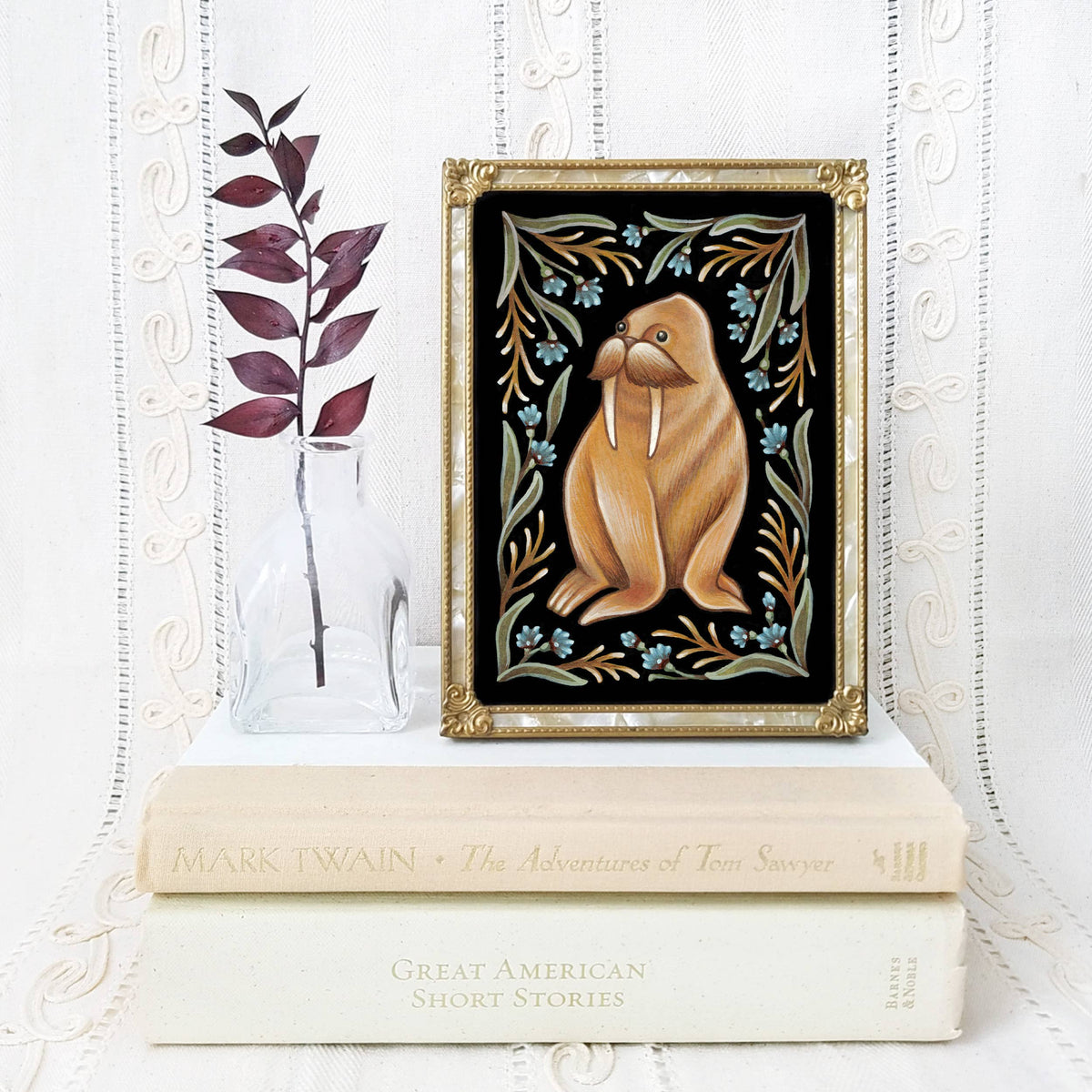 The Faina Lorah *Retiring* Walrus Folk Art Print (multiple sizes) features a walrus with floral patterns, displayed above stacked books beside a clear vase of dried burgundy leaves, set against a white textured background.