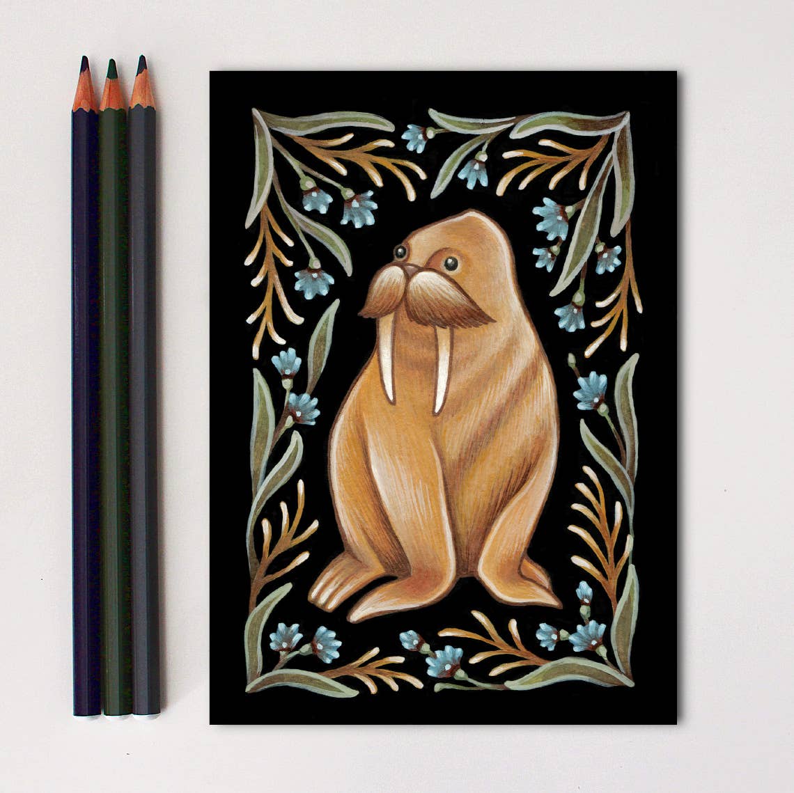 *Retiring* Walrus Folk Art Print by Faina Lorah features a brown walrus with tusks, green leaves, and blue flowers on black. Shown with three colored pencils on a white surface. Available in multiple sizes.