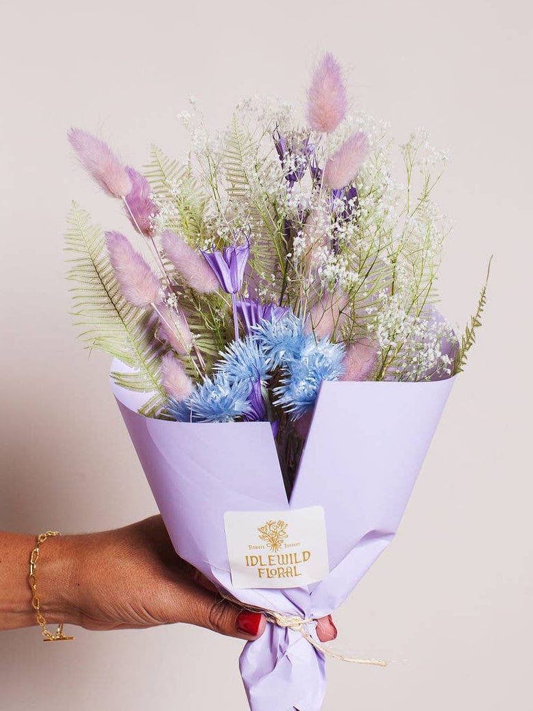 A hand holds the Farmhouse Dried Flower Bouquet by Idlewild Floral Co., featuring pastel preserved blooms in light purple, pink, and blue, wrapped in lavender paper with a branded label.