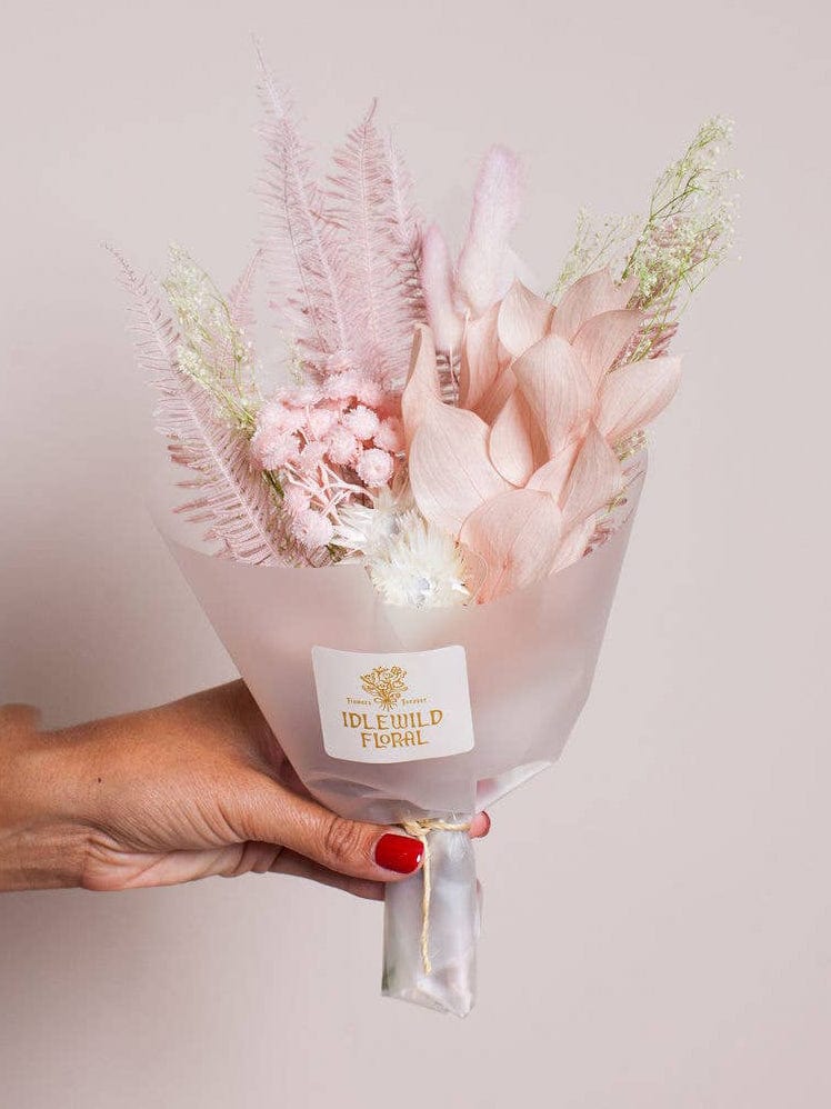 A hand holds the Lark Valentine Bouquet by Idlewild Floral Co., featuring pastel pink dried and preserved flowers with delicate foliage, wrapped in frosted paper against a pale background.