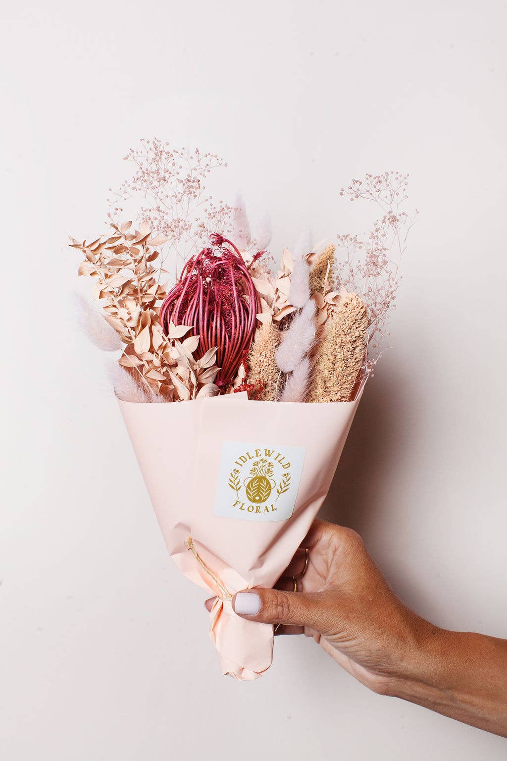 A hand holds Idlewild Floral Co.’s Strawberry Dried Flower Bouquet—petite pink and beige blooms wrapped in pale pink paper with a branded label—against a plain light background.