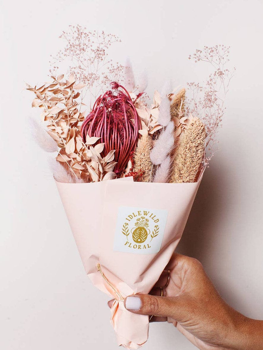 A hand holds the Strawberry Dried Flower Bouquet from Idlewild Floral Co., featuring dried blooms in pink, beige, and red tones, wrapped in pale pink paper with a gold brand label against an off-white background.