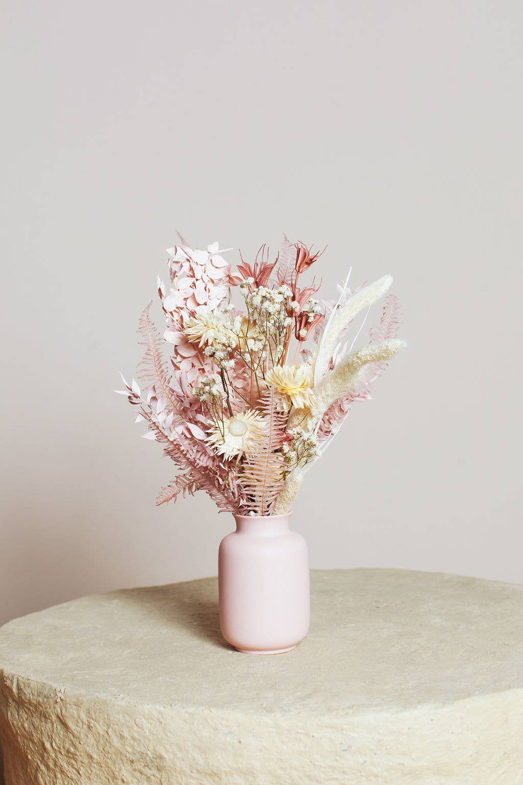 The Lark Valentine Bouquet by Idlewild Floral Co. features preserved pastel flowers and foliage arranged in a pink vase, displayed on a round, textured beige surface with a soft, light background.