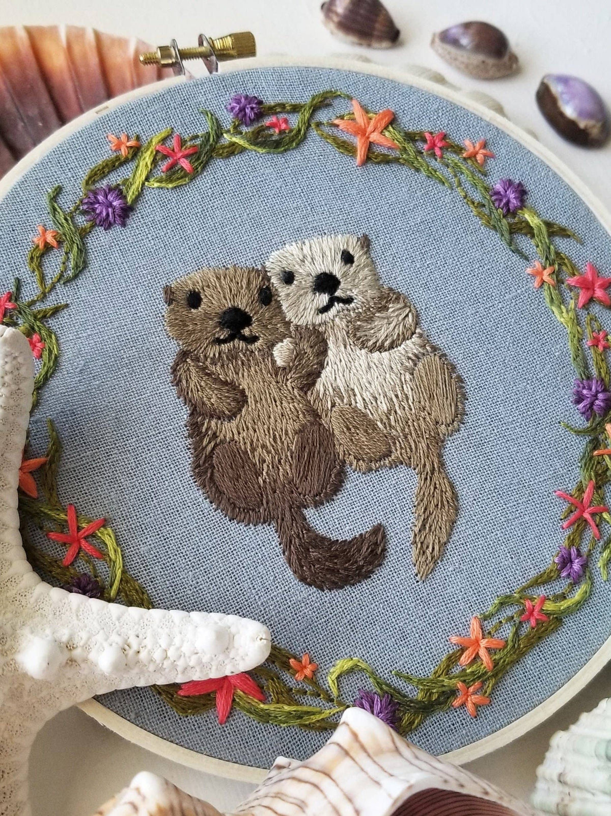 The *Late May Pre-Order* 6 Otterly Adorable Embroidery Kit by Jessica Long Embroidery features two otters holding hands with a floral and seaweed border—perfect for wall art or as a handmade gift.
