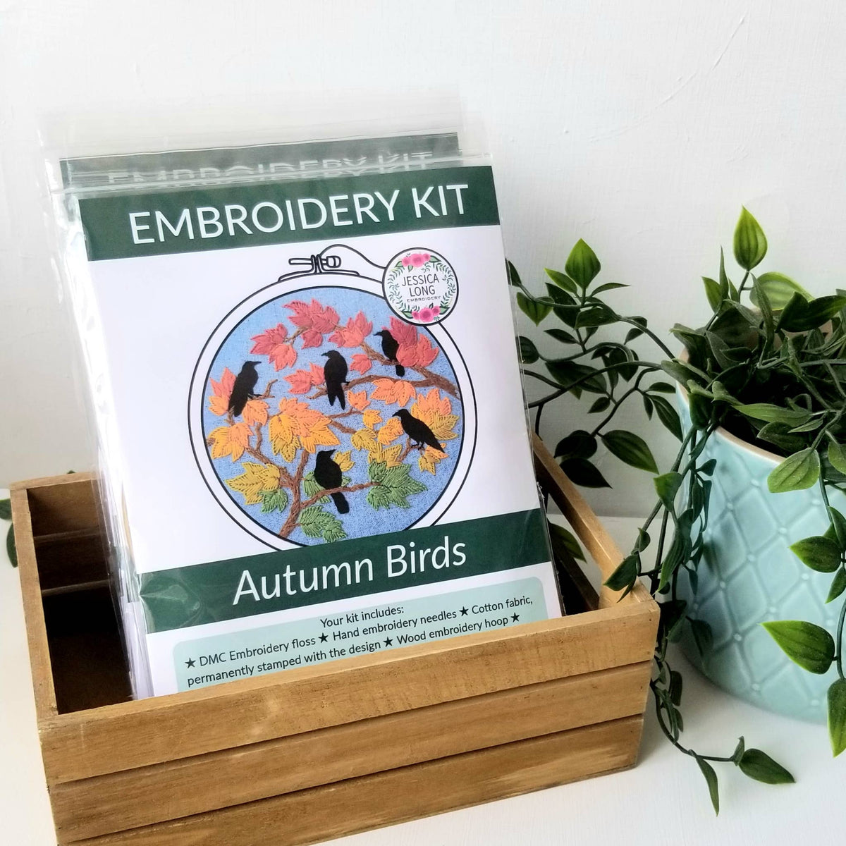The 6 Beginner Autumn Birds Embroidery Kit by Jessica Long Embroidery comes in a wooden box beside a green potted plant, featuring black birds on bright fall leaves and listing all included supplies on the packaging.