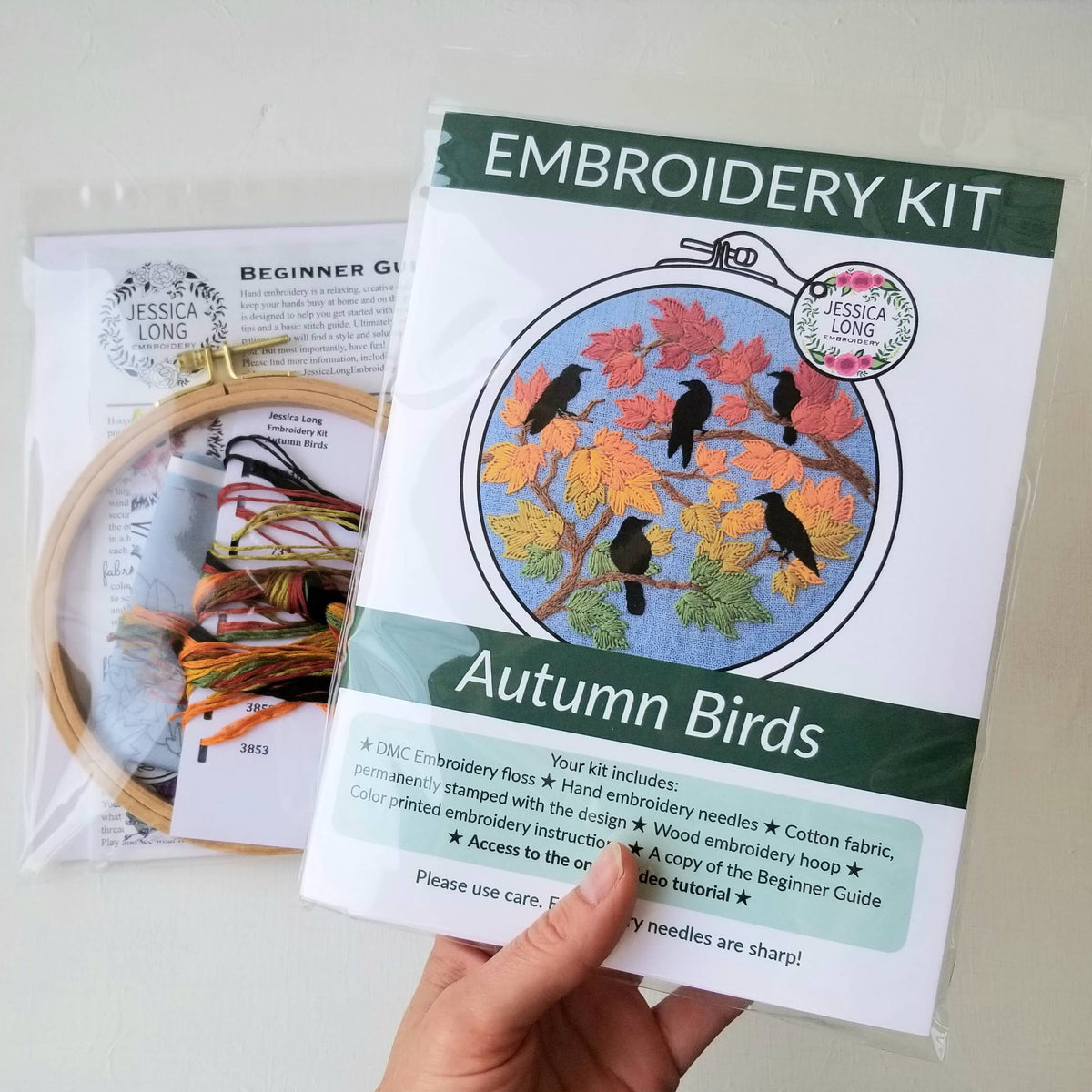 A hand holds the Jessica Long Embroidery 6 Beginner Autumn Birds Embroidery Kit with a pattern of black birds on fall leaves. Beside it is a package containing hoops, threads, needles, and an instruction guide.