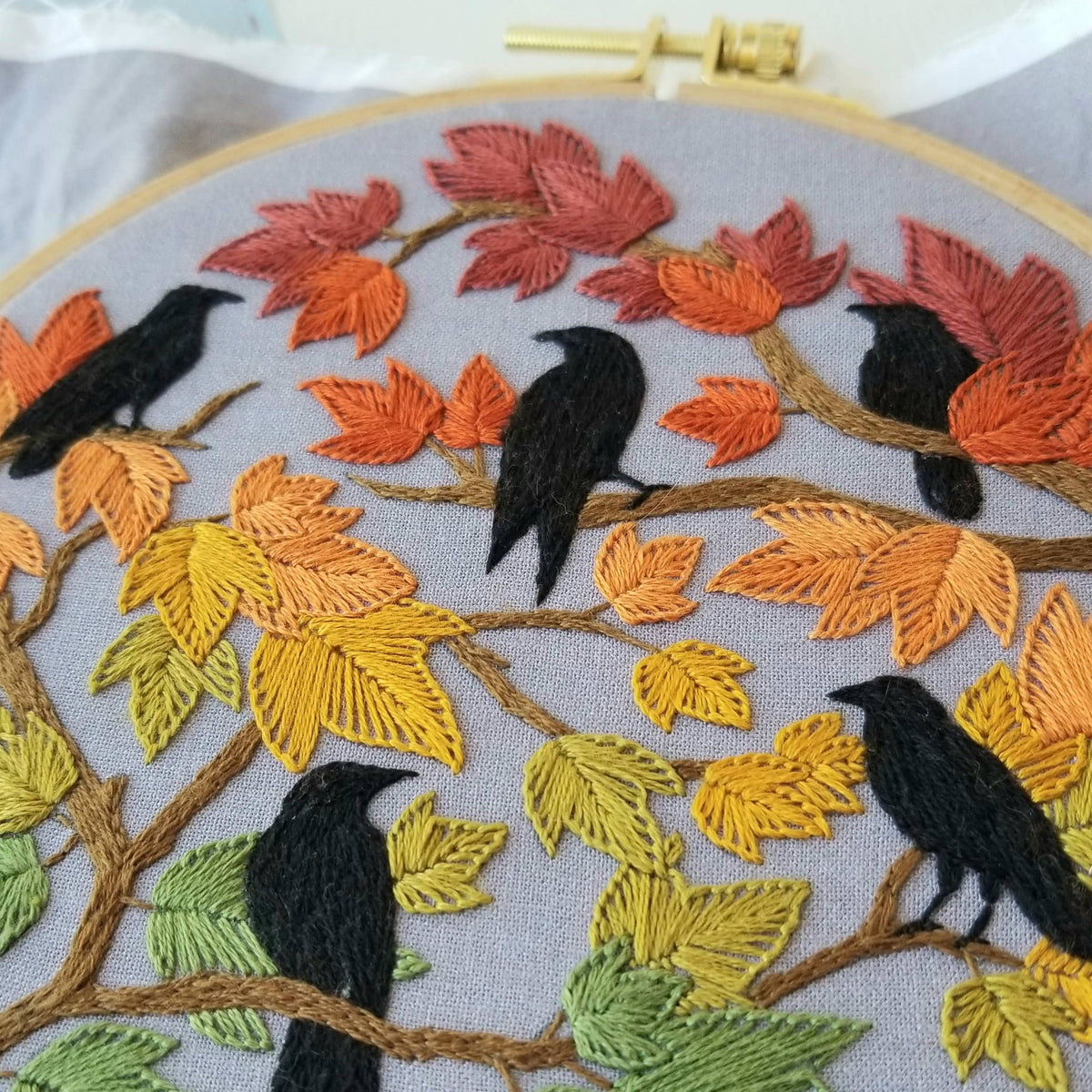 Jessica Long Embroiderys 6 Beginner Autumn Birds Embroidery Kit features five black crows on autumn branches with green, yellow, orange, and red leaves on gray fabric—perfect for a fall leaves craft or beginner embroidery project.