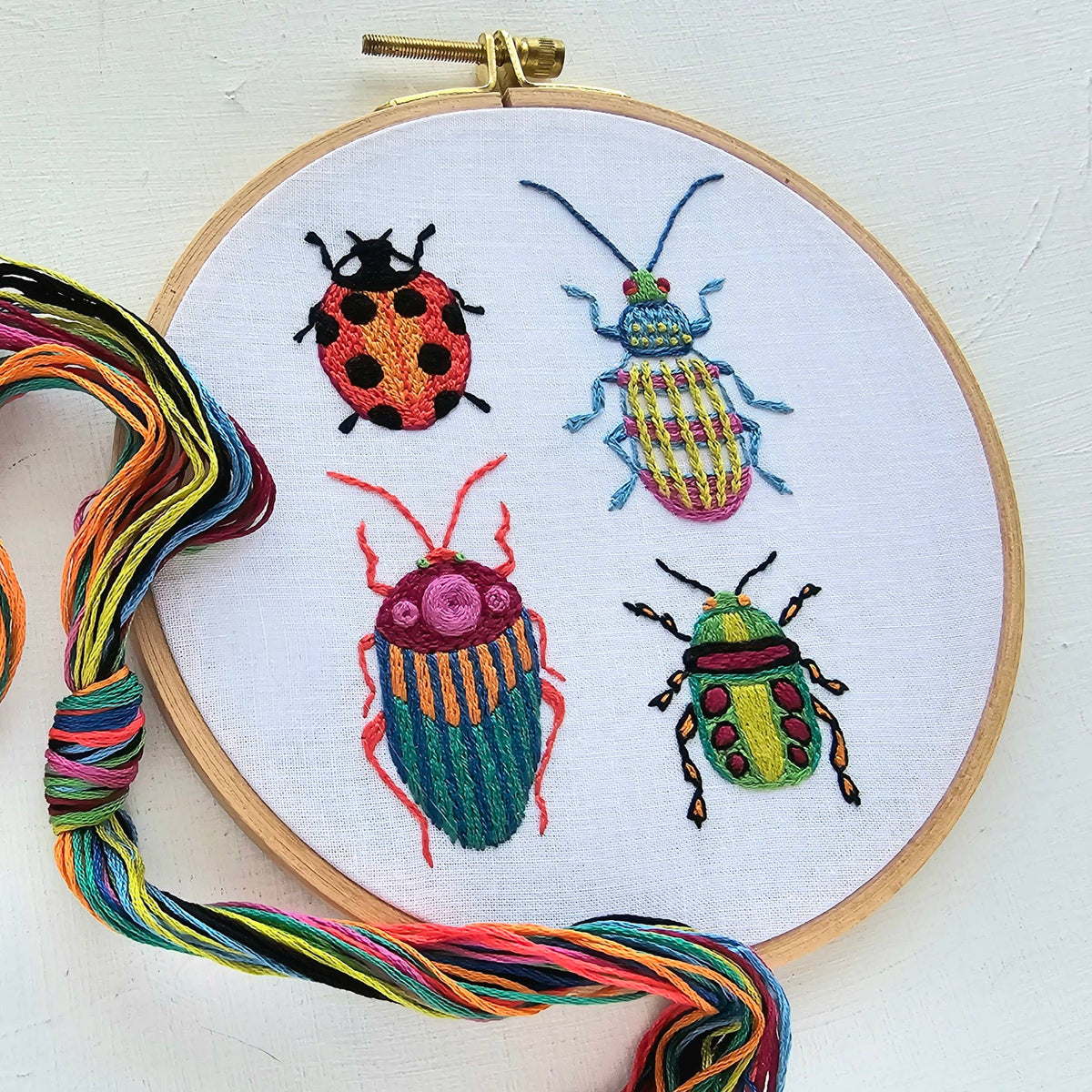 The 6 Beginner Beetle Collection Embroidery Kit by Jessica Long Embroidery features four uniquely patterned, colorful beetles on white fabric in a wooden hoop, plus multicolored threads—perfect for beginners or as a beetle sampler.