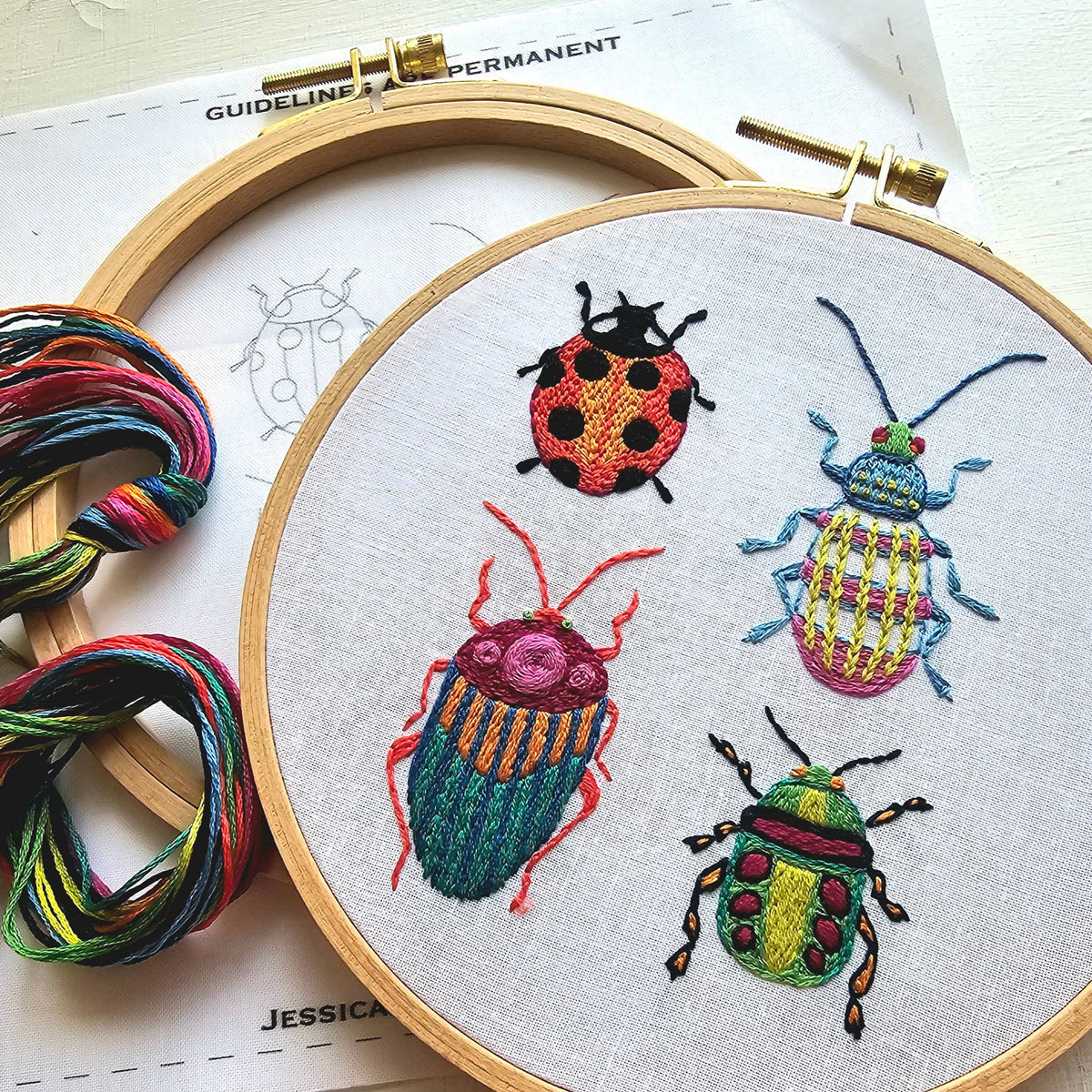 The Jessica Long Embroidery 6 Beginner Beetle Collection Embroidery Kit features a wooden hoop with four vibrant embroidered beetles on white fabric, multicolored floss, and a beetle sampler outline—perfect for beginners.