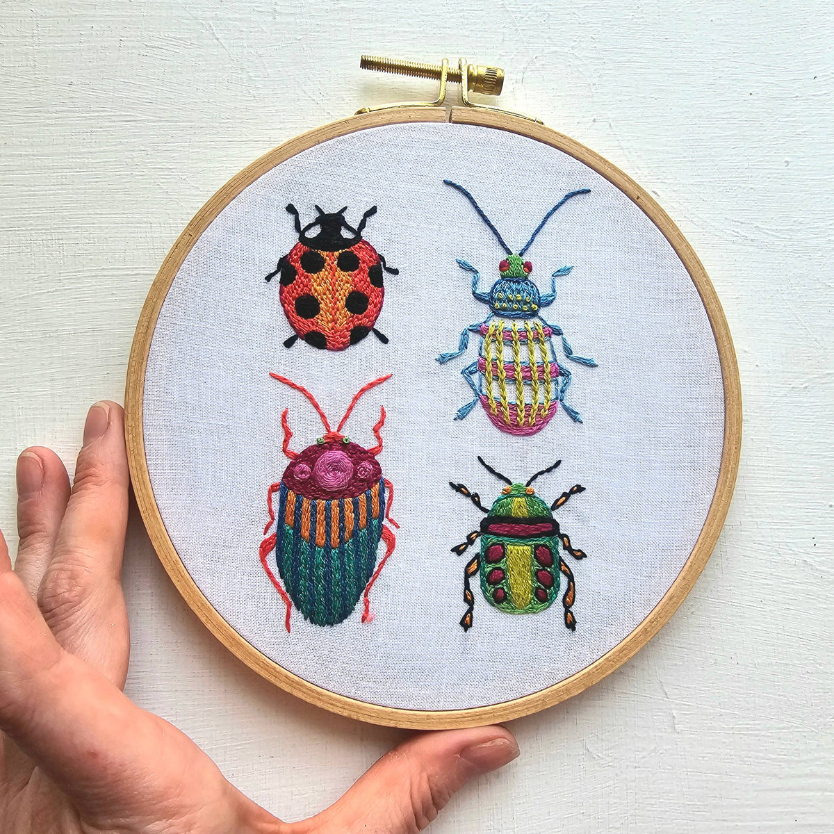 A hand displays the Jessica Long Embroidery 6 Beginner Beetle Collection Embroidery Kit, featuring a ladybug and three uniquely patterned beetles on white fabric—ideal as a beginner sampler or kit.