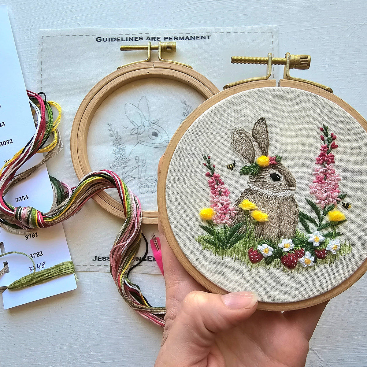 A hand holds Jessica Long Embroidery’s *Late May Pre-Order* 4 Berry Patch Bunny Embroidery Craft Kit, showing a rabbit in flowers and greenery made with thread painting; a color chart and pattern sketch rest on the light surface nearby.
