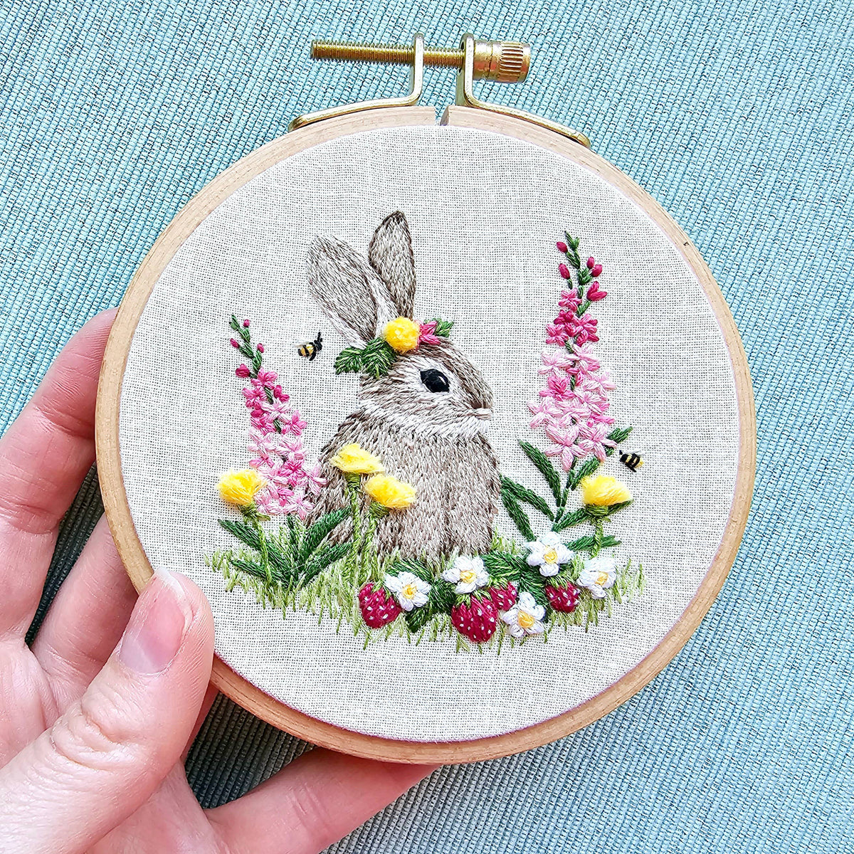A hand displays a finished 4 Berry Patch Bunny from Jessica Long Embroiderys *Late May Pre-Order* kit, featuring a gray rabbit with floral crown, surrounded by flowers, grass, strawberries, and bees—stitched on linen using thread painting.
