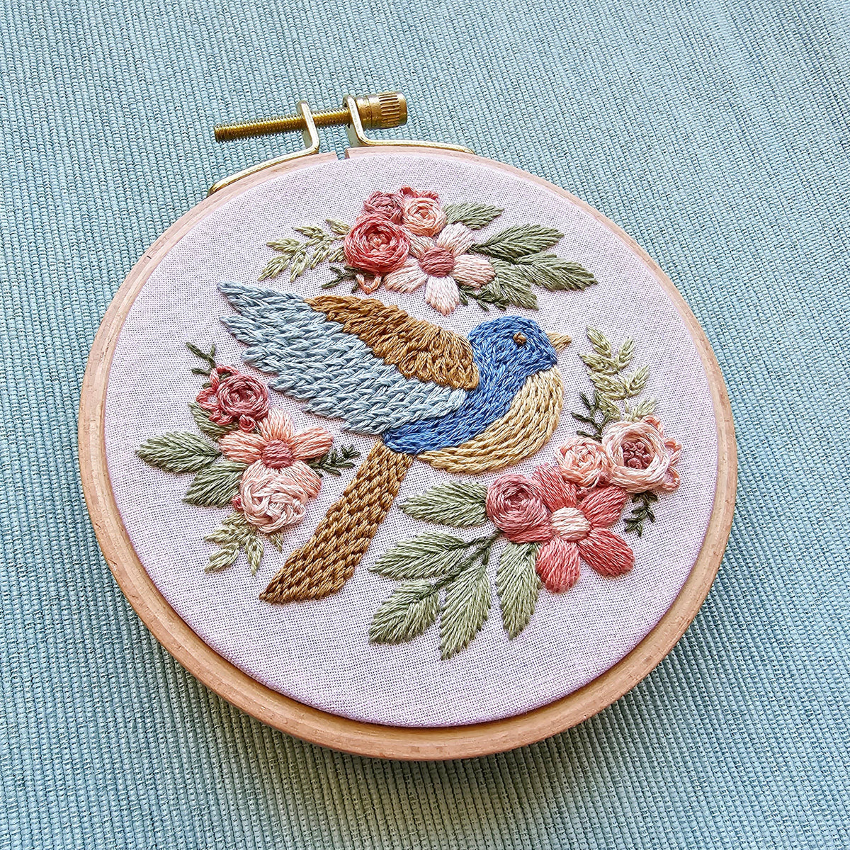 The 4 Beginner Bluebird Sampler Embroidery Kit by Jessica Long Embroidery features a blue and brown bird among pink roses and green leaves, stitched on light fabric—a charming starter set shown on a light blue textured surface.