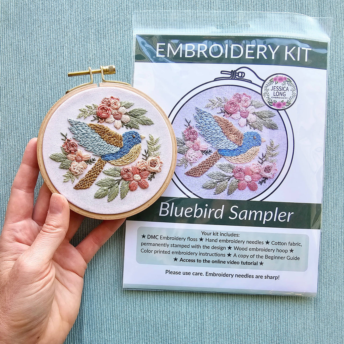 A hand holds a finished bluebird sampler in a hoop next to the Jessica Long Embroidery 4 Beginner Bluebird Sampler Embroidery Kit, with its white package displaying the same floral design and included materials list.