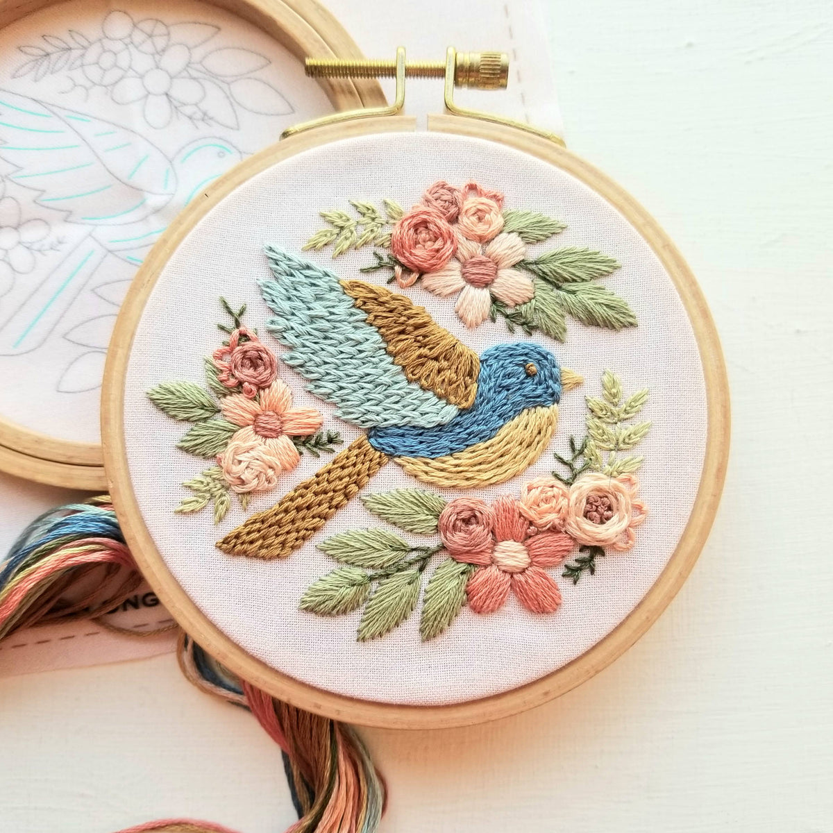 The Jessica Long Embroidery 4 Beginner Bluebird Sampler Embroidery Kit features a colorful bird among pink flowers and green leaves on white fabric, with included floss and a partially visible pattern for easy stitching.