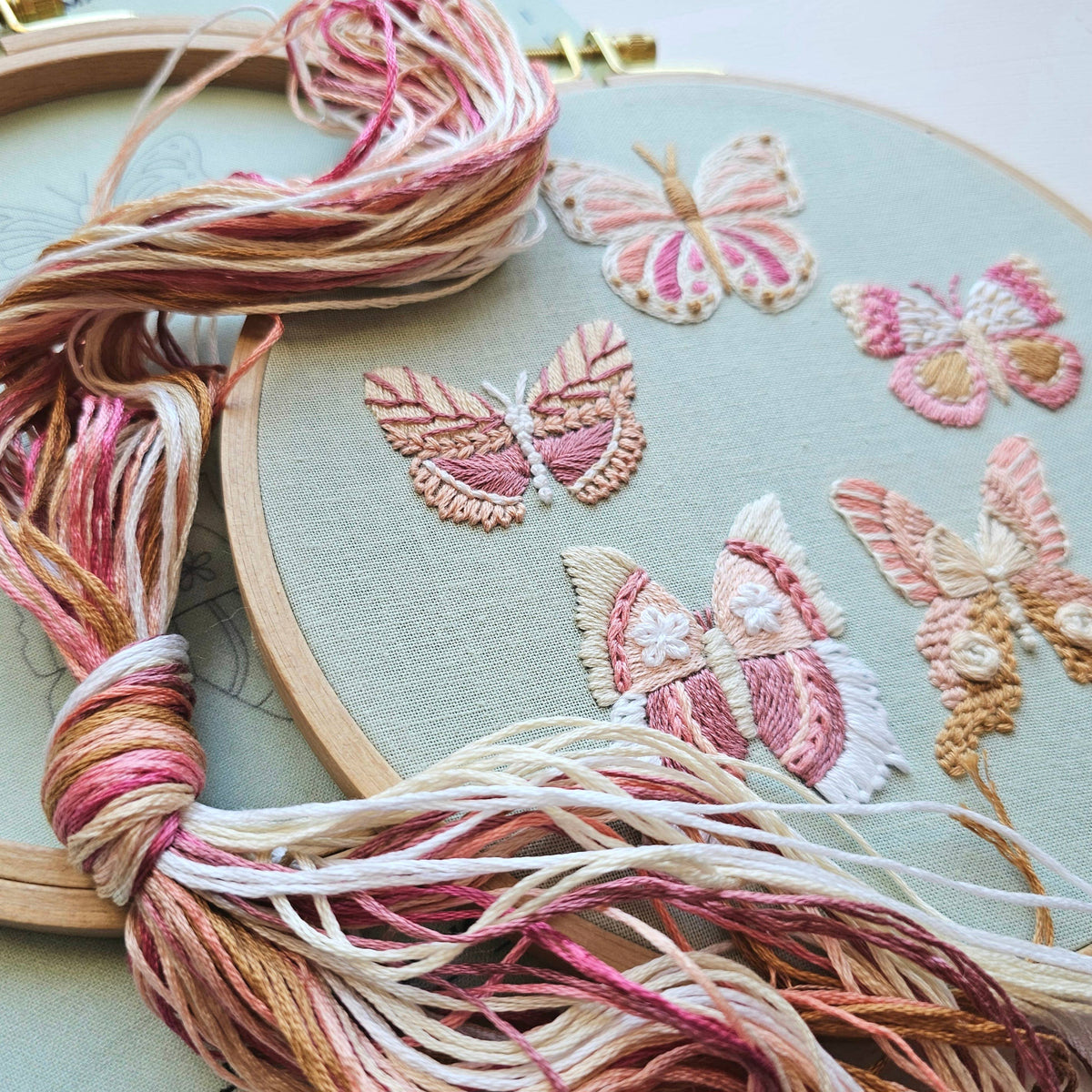 The Jessica Long Embroidery 6 Butterfly Embroidery Kit features a close-up of embroidered butterflies in pink, cream, and brown with matching threads and loose floss beside the hoop—ideal for beginner embroidery projects.