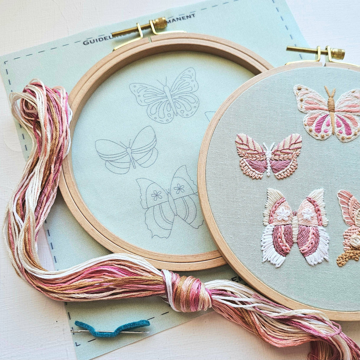 The 6 Butterfly Embroidery Kit by Jessica Long Embroidery features wooden hoops displaying butterfly sketches, partially stitched in pink and white thread, with multicolored floss and a blue felt butterfly to complete this beginner-friendly set.
