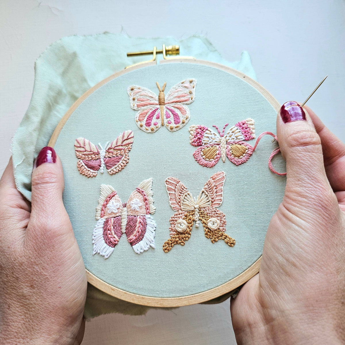 A pair of hands holds a 6 Butterfly Embroidery Kit by Jessica Long Embroidery, featuring detailed butterfly designs in pink, white, and brown. One hand uses a needle to stitch the pattern from this beginner-friendly kit.