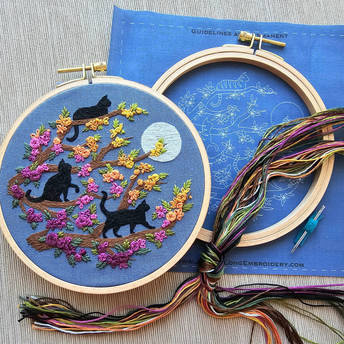 The Jessica Long Embroidery 6 Beginner Black Cats & Full Moon Embroidery Kit features two hoops: one with black cats on a flowering tree under a full moon, and a beginner’s hoop, all displayed on a textured beige surface.