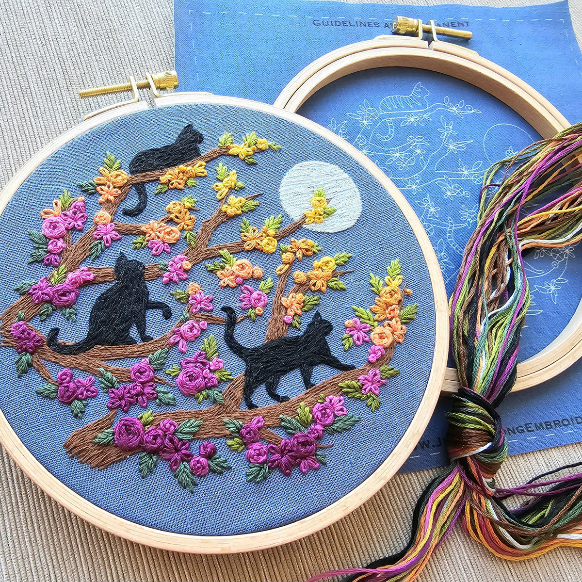 The Jessica Long Embroidery 6 Beginner Black Cats & Full Moon Embroidery Kit features black cats on tree branches with pink and yellow flowers, a full moon, and a blue sky—perfect for beginner stitchers.