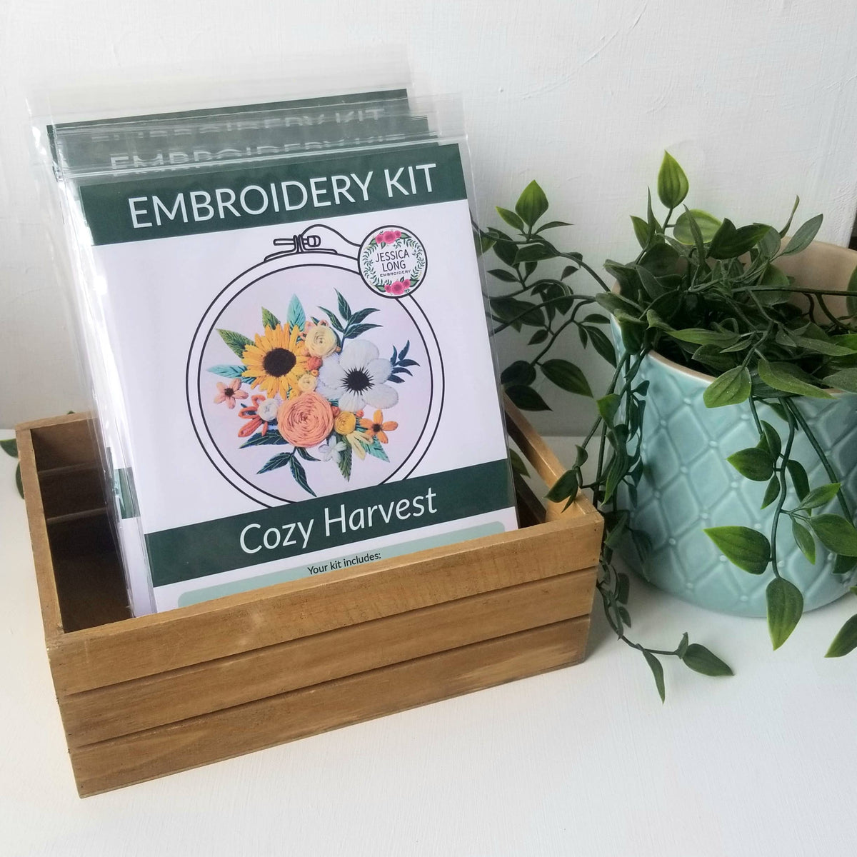 The 4 Beginner Cozy Harvest Embroidery Kit by Jessica Long Embroidery, featuring a floral design, rests in a wooden box beside a light blue textured plant pot with greenery—an ideal choice for new crafters.
