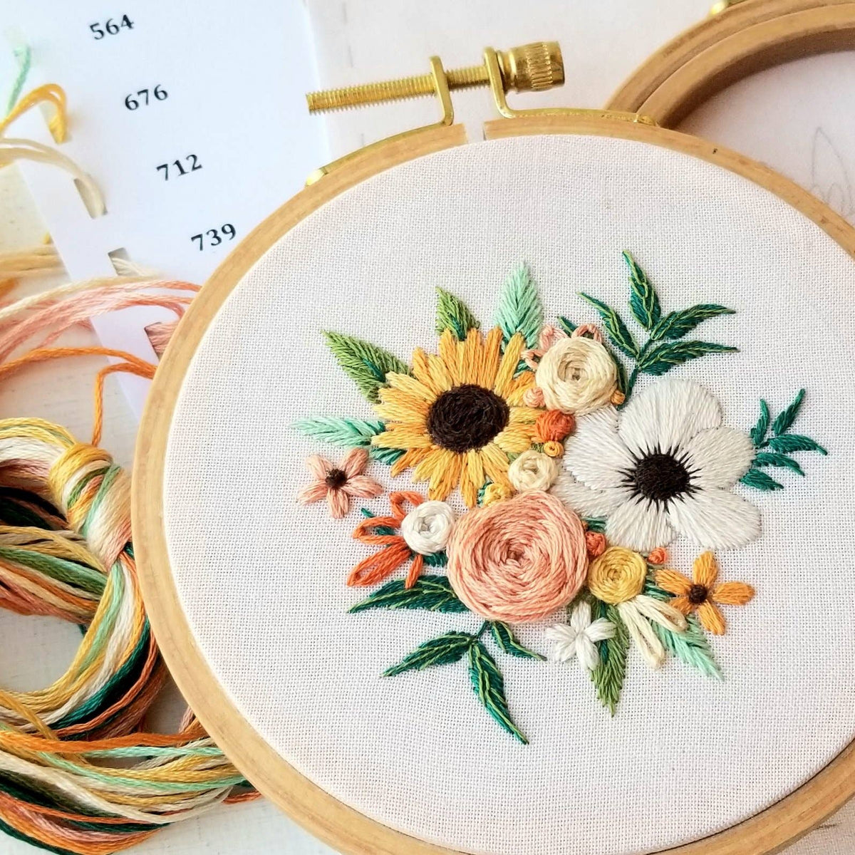 The 4 Beginner Cozy Harvest Embroidery Kit by Jessica Long Embroidery includes a hoop holding fabric with sunflower and floral designs, plus threads and a color card, making it perfect for beginners.