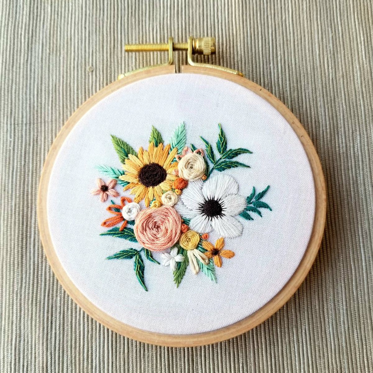 The 4 Beginner Cozy Harvest Embroidery Kit by Jessica Long Embroidery features a wooden hoop holding white fabric adorned with vibrant sunflowers, anemones, green leaves, and small blooms—perfect for beginners.