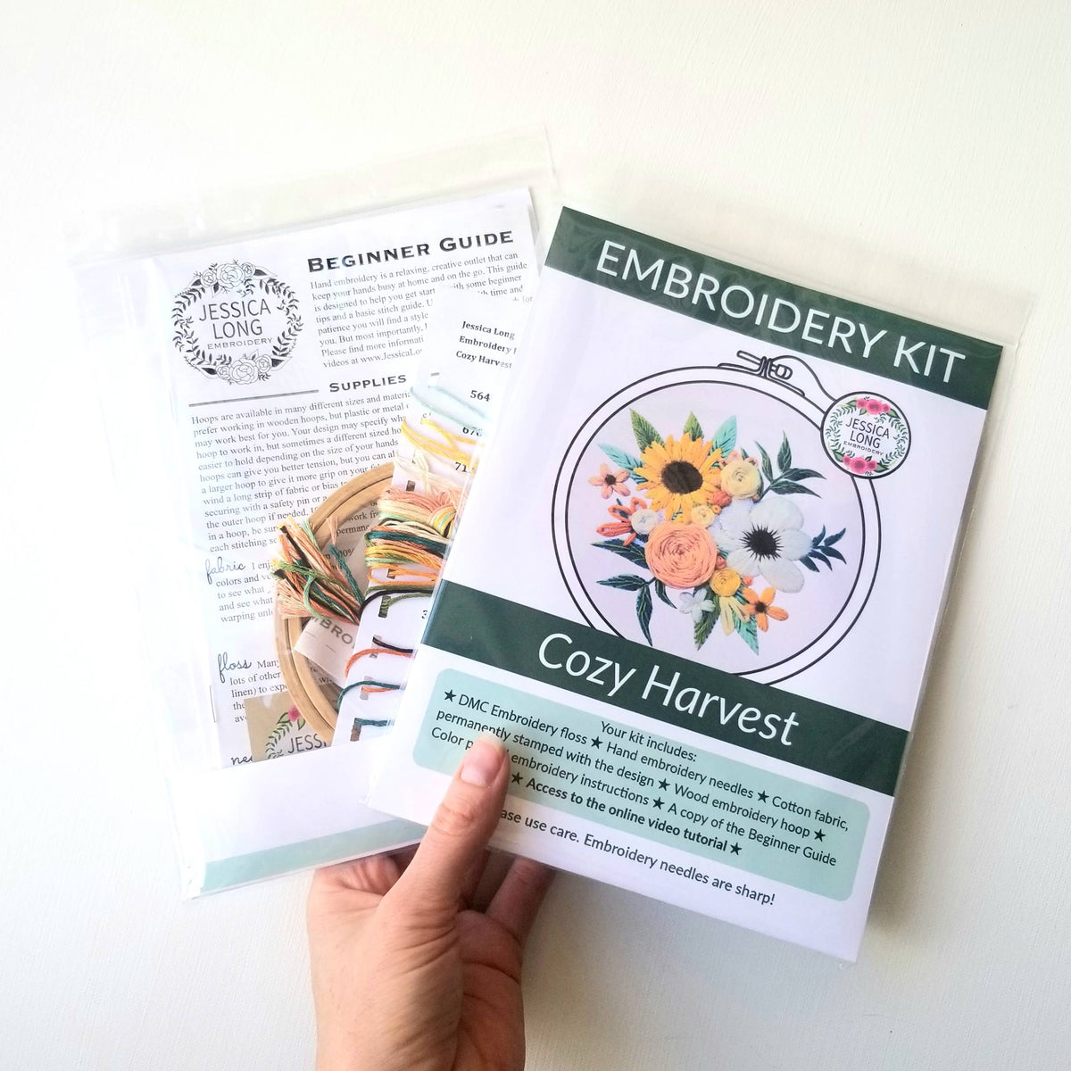 A hand holds Jessica Long Embroiderys 4 Beginner Cozy Harvest Embroidery Kit, featuring floral designs and a guide with floss and supplies, all neatly packed in clear packaging.