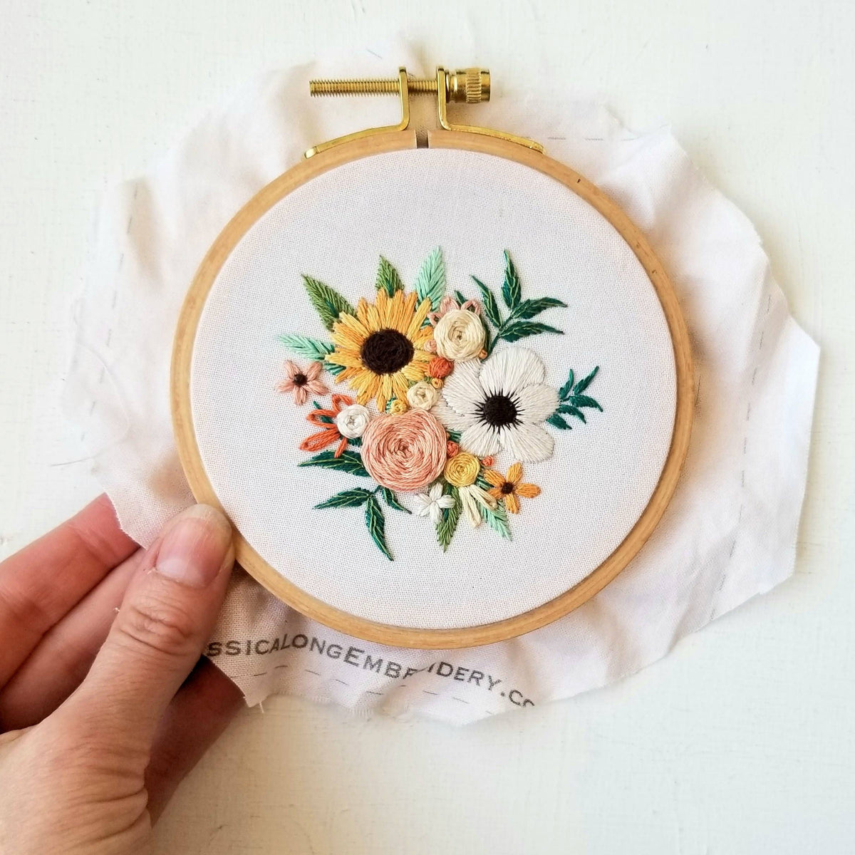 A hand holds the Jessica Long Embroidery 4 Beginner Cozy Harvest Embroidery Kit, featuring a floral design with sunflowers, roses, and colorful blooms stitched on white fabric.