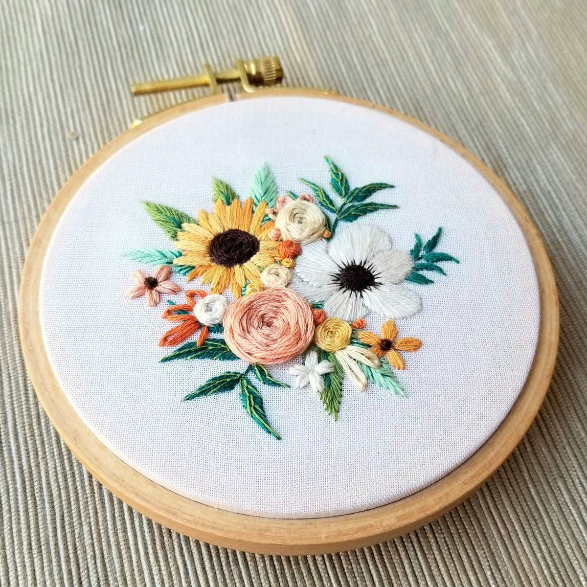 The 4 Beginner Cozy Harvest Embroidery Kit by Jessica Long Embroidery features a round wooden hoop displaying sunflowers, white and pink blooms, and small orange and yellow flowers against a light, textured background.