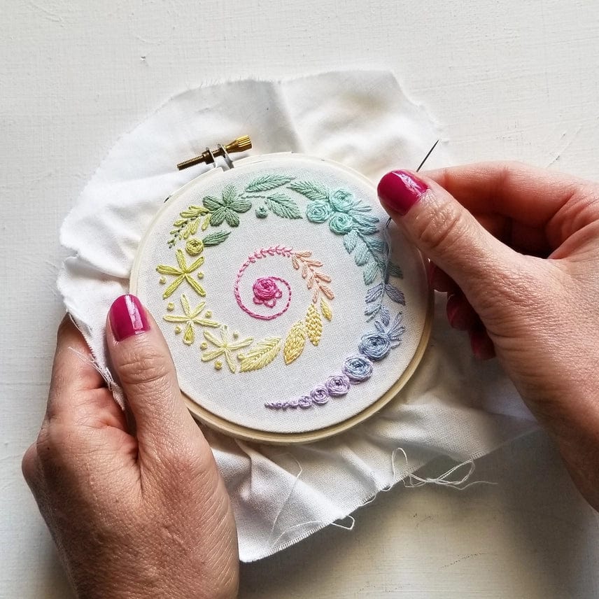 Two hands with pink nail polish hold the Jessica Long Embroidery 4 Beginner Ivory Spiral Sampler Embroidery Kit, stitching vibrant flowers and leaves on white fabric—a relaxing hobby for creative minds.