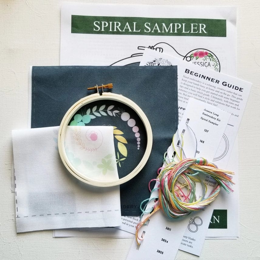 The 4 Beginner Ivory Spiral Sampler Embroidery Kit by Jessica Long Embroidery includes a wooden hoop, patterned ivory fabric, colorful threads, and instructions—perfect for beginners or as a relaxing hobby—displayed on a white surface.