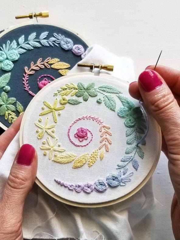 A person with pink nail polish works on pastel floral designs using the 4 Beginner Ivory Spiral Sampler Embroidery Kit by Jessica Long Embroidery, while a darker finished piece sits in the background, capturing this relaxing hobby.