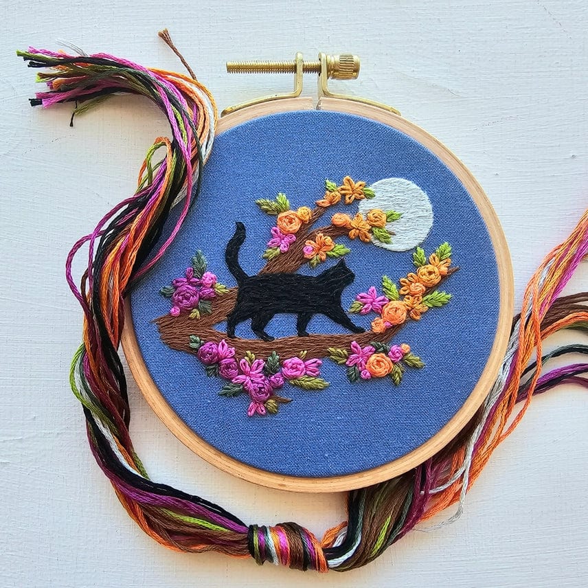 The Jessica Long Embroidery 4 Beginner Mini Black Cats & Full Moon Embroidery Kit features a black cat on a tree branch with colorful flowers and a white moon on blue fabric—multi-colored threads included, perfect for beginners.