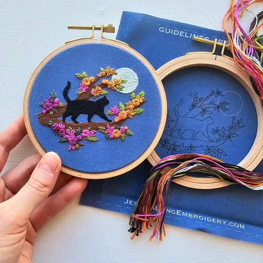A hand holds the Jessica Long Embroidery 4 Beginner Mini Black Cats & Full Moon Embroidery Kit, featuring a black cat on a branch with vibrant flowers and a full moon, while an unfinished kit and colorful threads inspire creativity nearby.