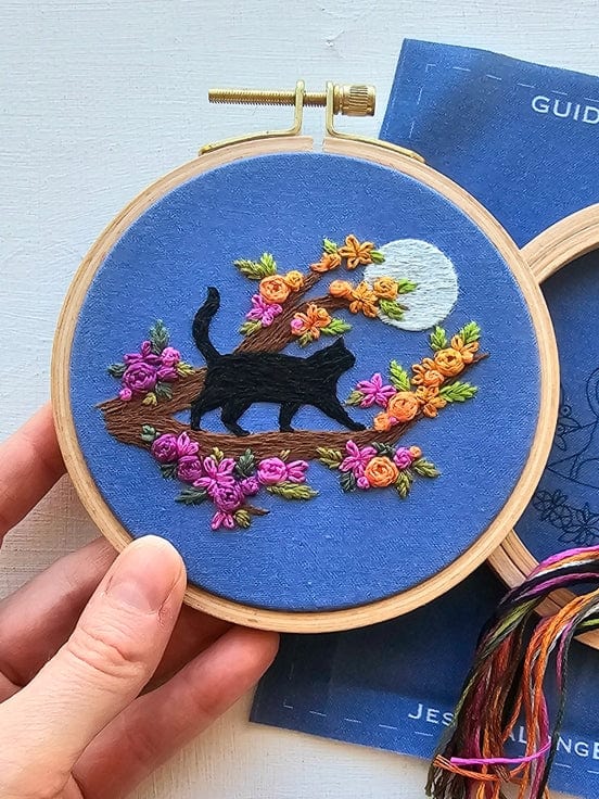 A hand holds the Jessica Long Embroidery 4 Beginner Mini Black Cats & Full Moon Kit, featuring a stitched black cat on a branch with colorful flowers, leaves, and a moon on blue fabric—perfect for embroidery beginners.
