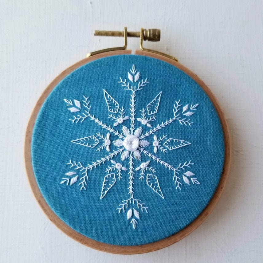 The Jessica Long Embroidery 4 Beginner Mini Snowflake Sampler Kit features teal fabric in a wooden hoop with a detailed white snowflake embroidered in various stitch patterns for added texture and depth.