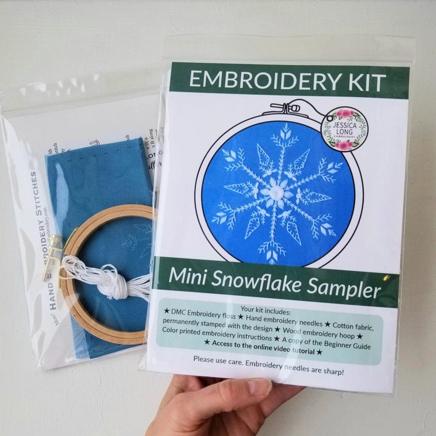 A hand holds the Jessica Long Embroidery 4 Beginner Mini Snowflake Sampler Embroidery Kit, featuring blue fabric with a white snowflake, hoop, thread, and instructions—ideal as a stocking stuffer or for winter hoop art projects.