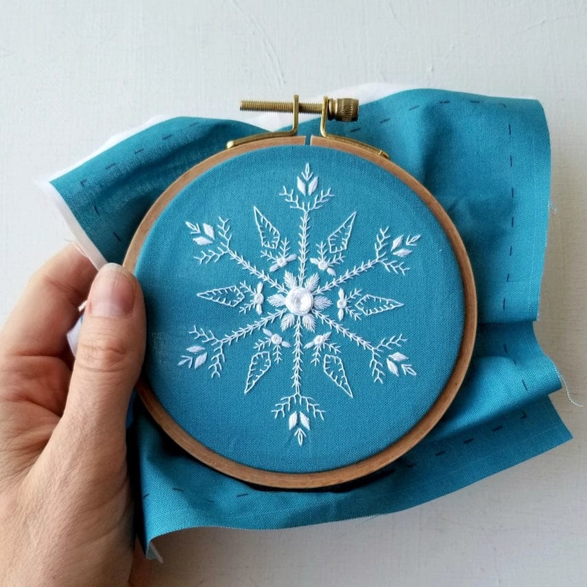 A hand holds the Jessica Long Embroidery 4 Beginner Mini Snowflake Sampler Kit, featuring a wooden hoop with turquoise fabric and a detailed white embroidered snowflake—a charming winter embroidery project.