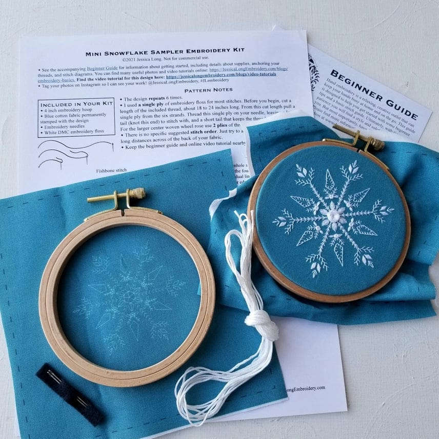 The Jessica Long Embroidery 4 Beginner Mini Snowflake Sampler Kit includes two blue fabric squares (one blank, one in a hoop with a white snowflake), white & dark thread, needle, and instructions—perfect for DIY embroidery or a stocking stuffer gift.