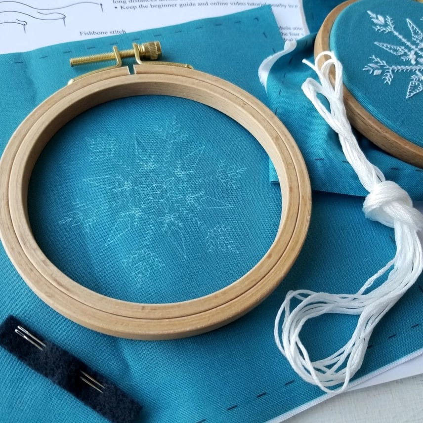 The 4 Beginner Mini Snowflake Sampler Embroidery Kit by Jessica Long Embroidery includes a wooden hoop, teal snowflake fabric, white thread, extra fabric, black felt, and instructions—ideal for winter DIY art or as a cute stocking stuffer.