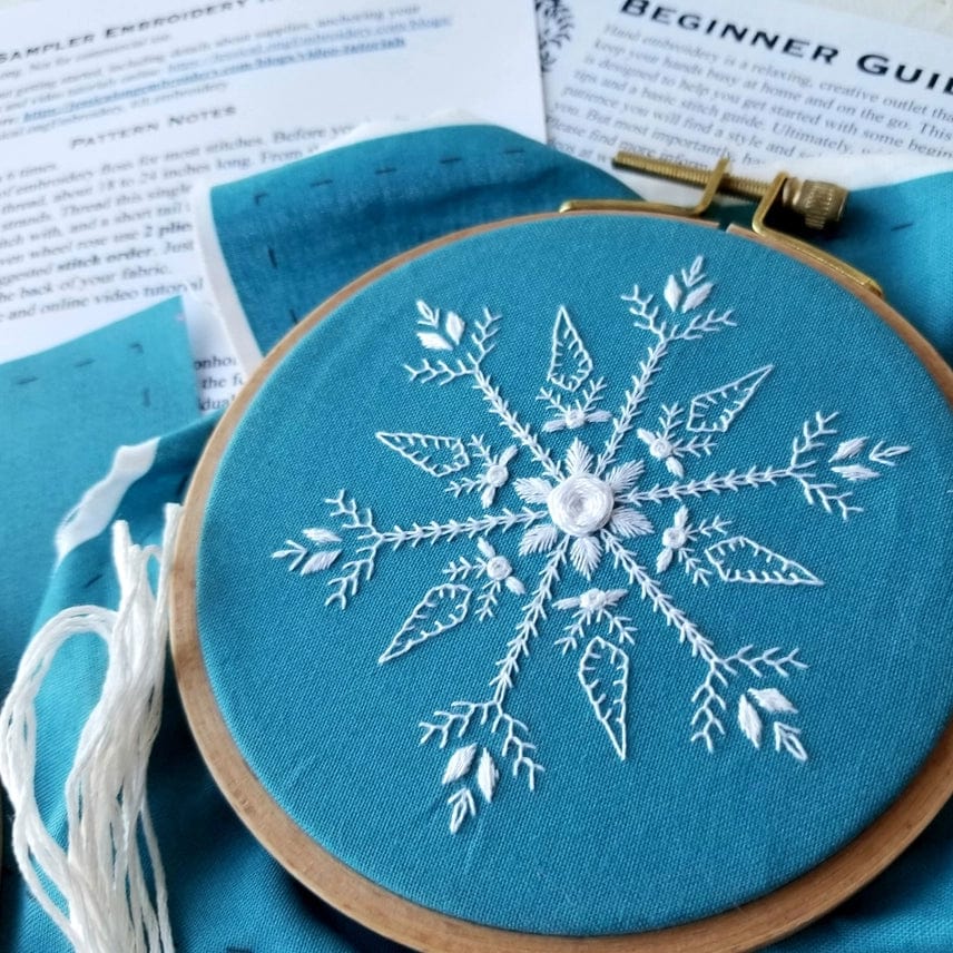 The Jessica Long Embroidery 4 Beginner Mini Snowflake Sampler Kit includes a round hoop, teal fabric with a white snowflake design, thread, and instructions—perfect for winter art or as a stocking stuffer gift.