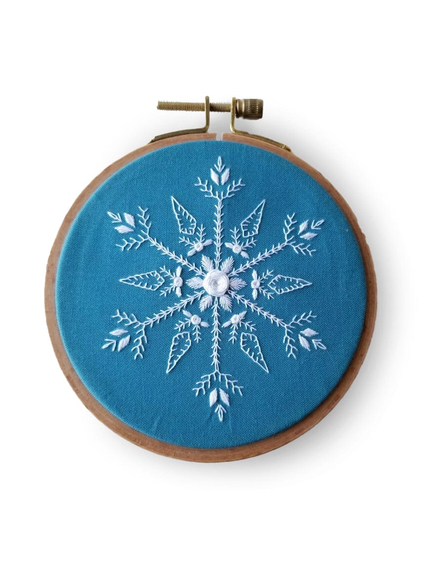 The 4 Beginner Mini Snowflake Sampler Embroidery Kit by Jessica Long Embroidery features a detailed white snowflake on blue fabric in a wooden hoop—ideal for beginners and makes a great stocking stuffer gift.